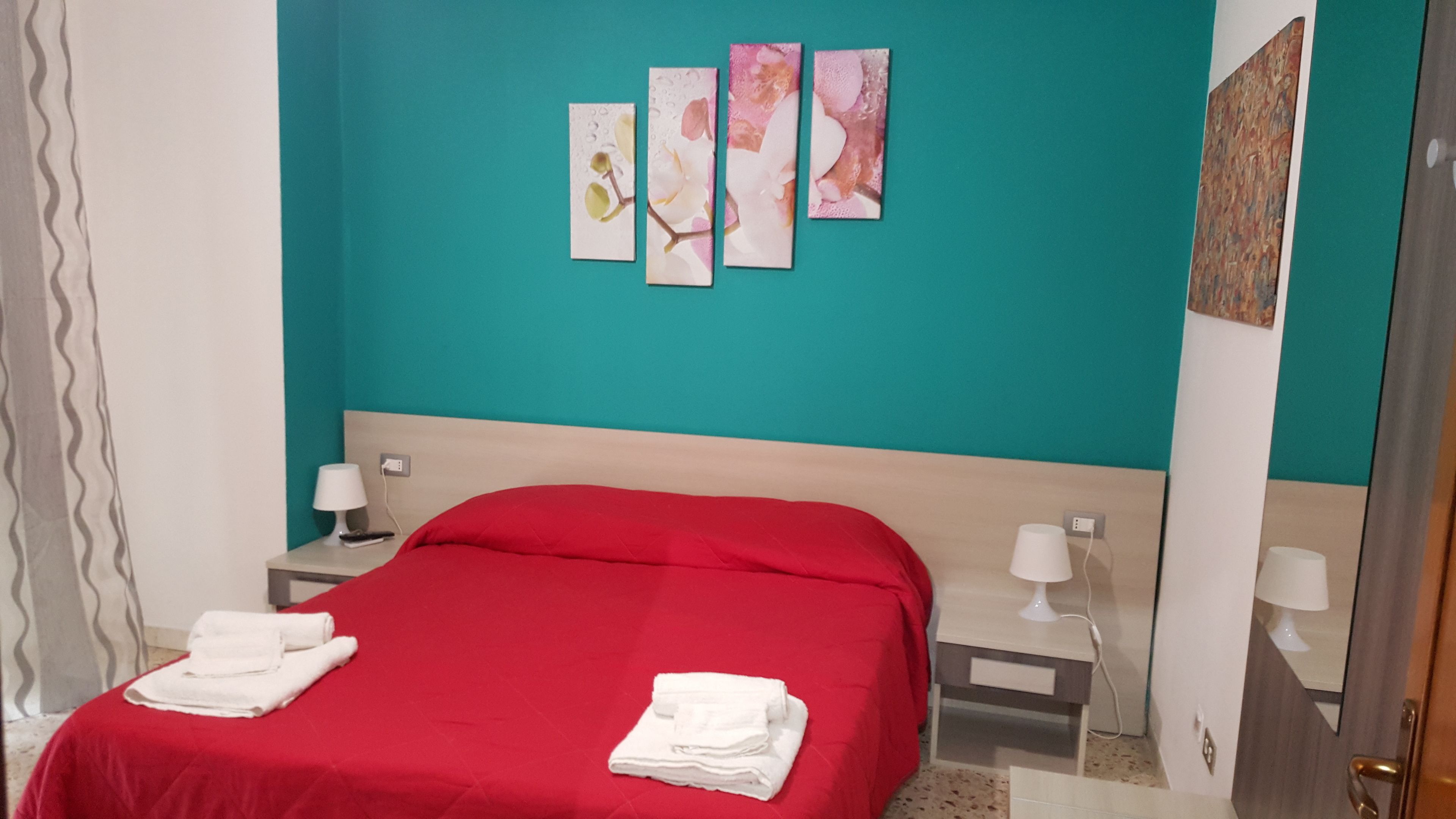 A bedroom with a teal wall, a red bedspread, and a white headboard. There are two bedside tables with lamps and a painting above the bed.