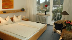 A hotel room with a bed, a bedside table with a lamp, a chair, a small table with a vase of flowers, a window with a view of trees and buildings, and a radiator. - Hotel Ebnet Garni (Lindau)