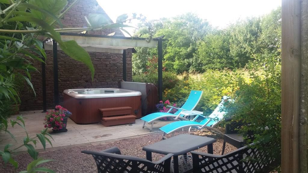 Un jacuzzi bajo una pérgola, rodeado de una terraza de madera y mobiliario de jardín.