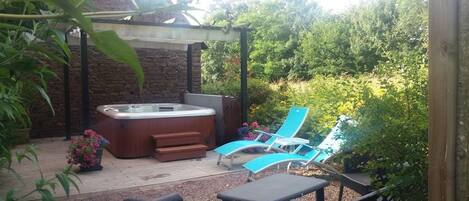 Un jacuzzi bajo una pérgola, rodeado de una terraza de madera y mobiliario de jardín.