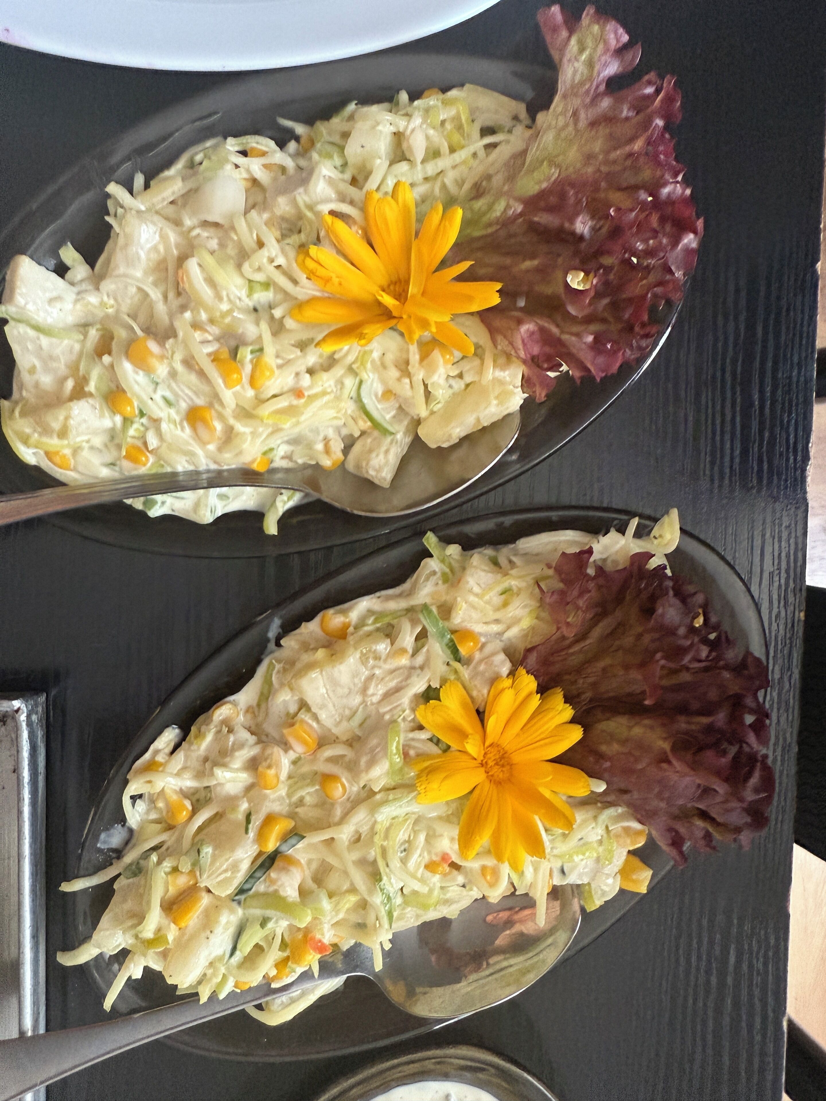 Two plates of coleslaw with corn and a yellow flower garnish, served on a black table.