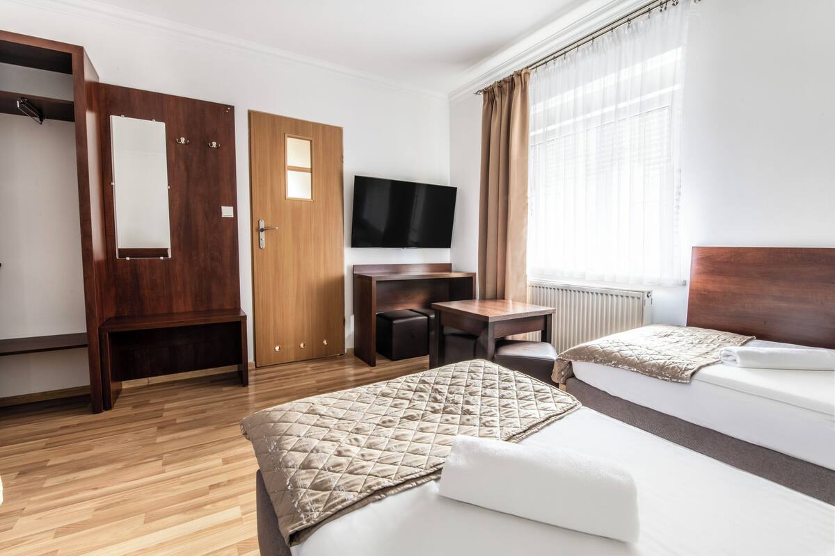 A hotel room with a wooden wardrobe, a desk with a chair, a television, and two beds with a quilted blanket.