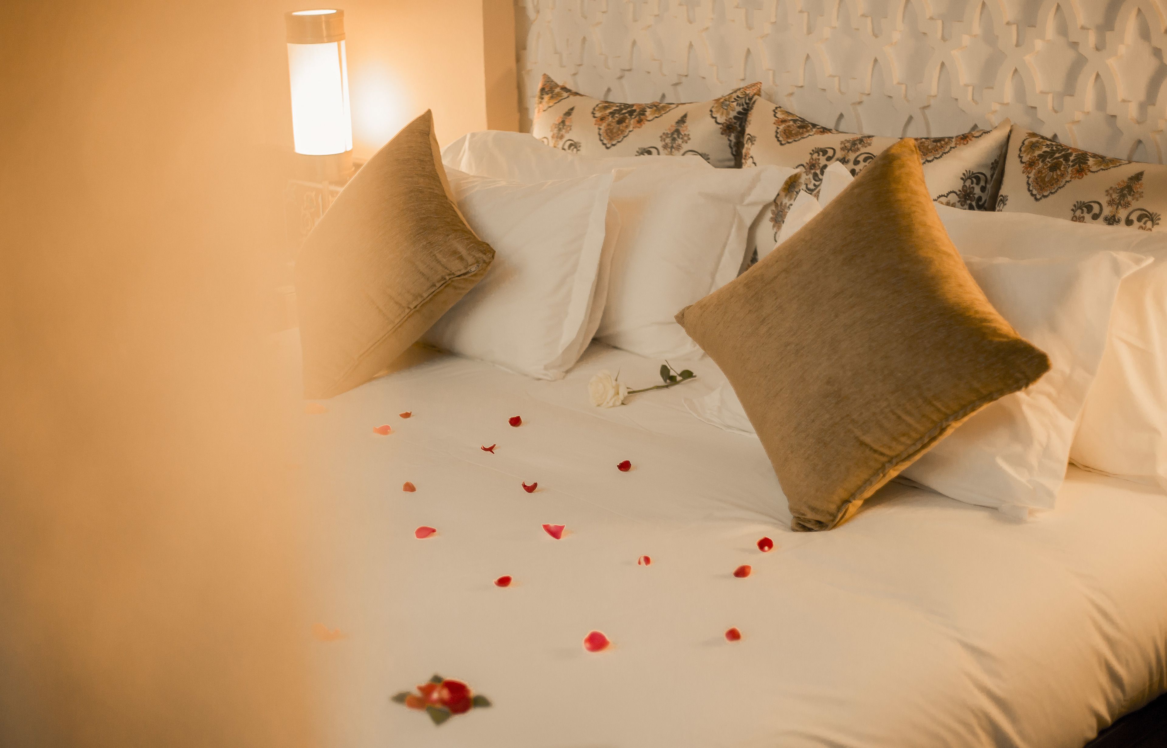 A neatly made bed with white and beige pillows, a decorative headboard, and rose petals scattered on the bed.