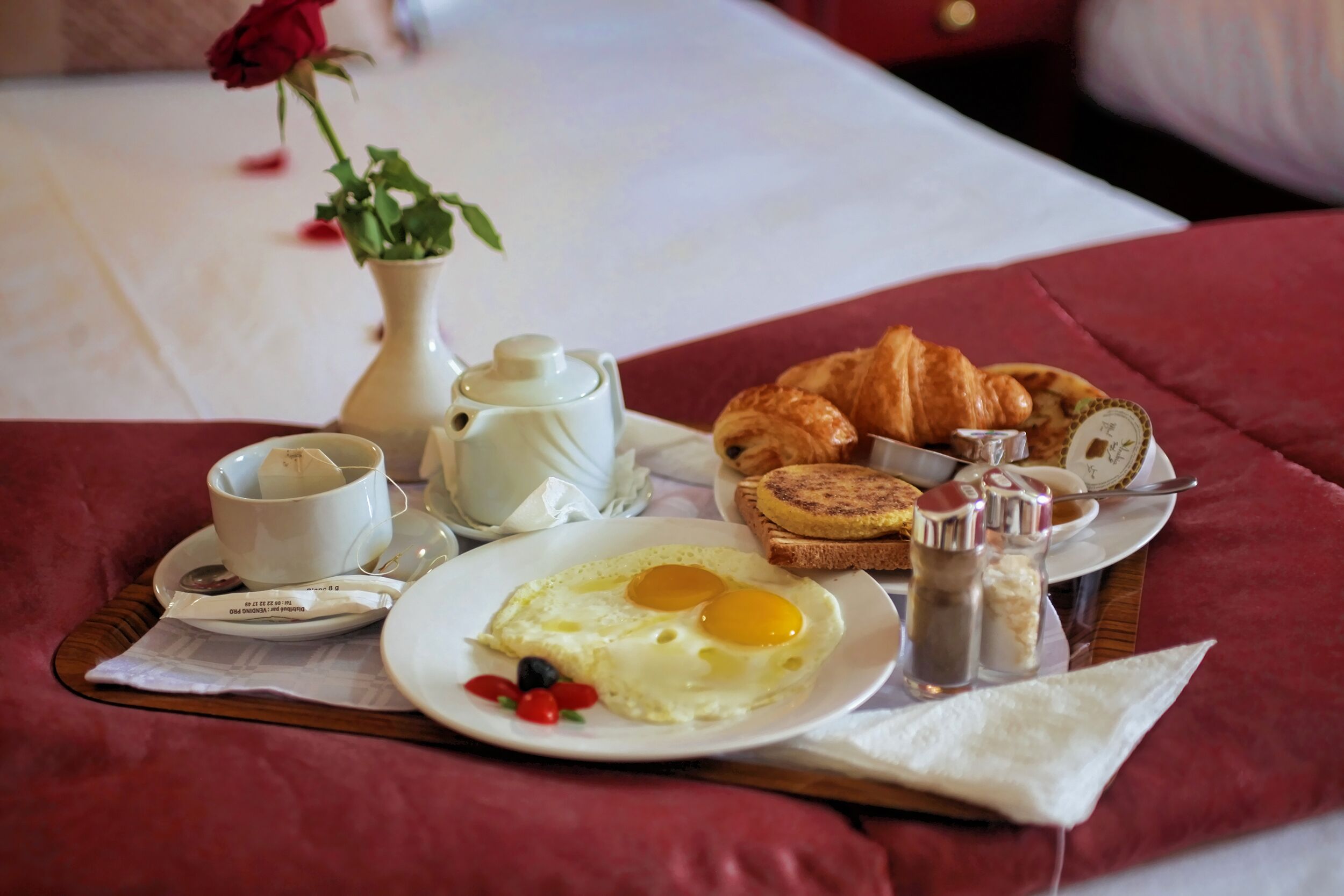A breakfast tray with eggs, toast, and a rose on a bed.