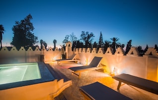 A rooftop pool area with lounge chairs and a hot tub, surrounded by a decorative wall and palm trees.