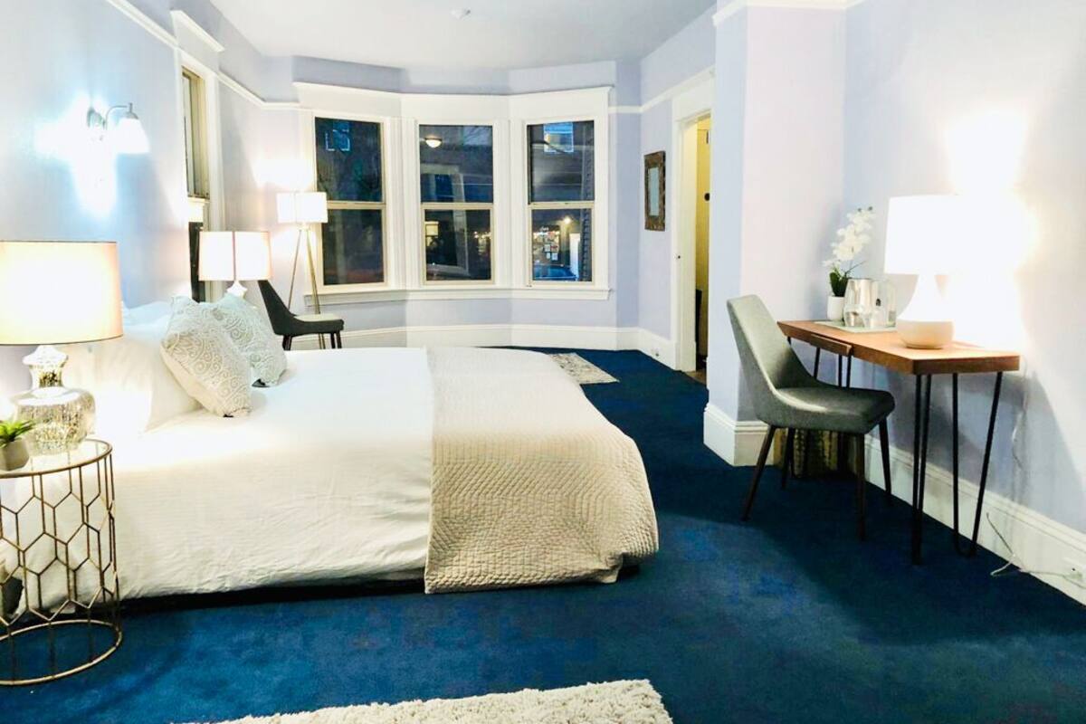 A bedroom with a large bed, a desk, and a chair. There is a window with a view of the outside.