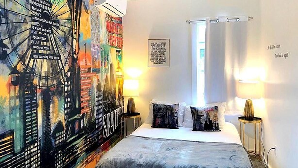 A hotel room with a large cityscape mural, a bed with two pillows, a nightstand with a lamp, and a window with a curtain. - The ART INN Seattle (Seattle)