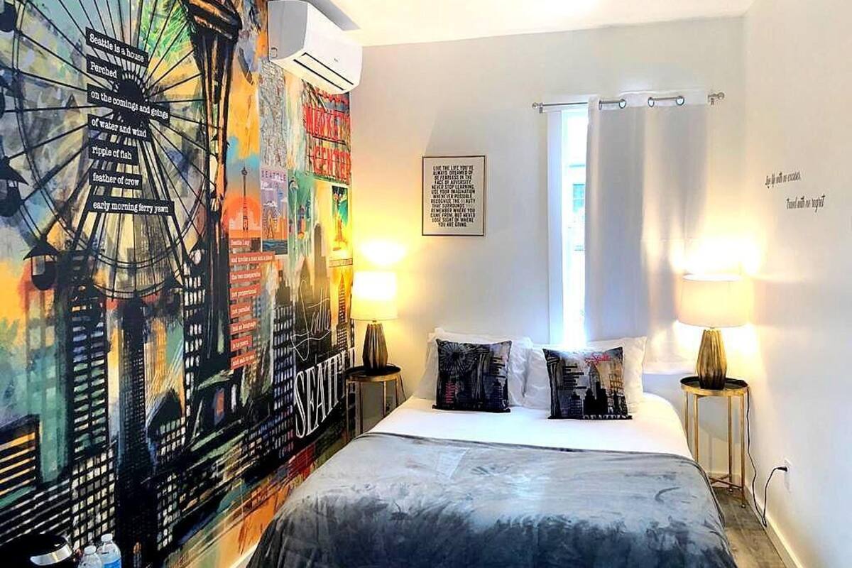 A hotel room with a large cityscape mural, a bed with two pillows, a nightstand with a lamp, and a window with a curtain.