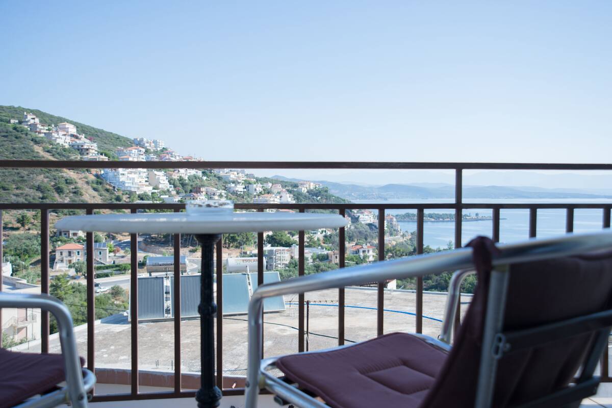 A balcony with two cushioned chairs and a small table, overlooking a coastal town with buildings and a body of water.