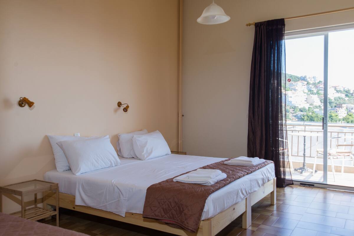 A bedroom with a bed, a small table, and a view of the city through a large window.
