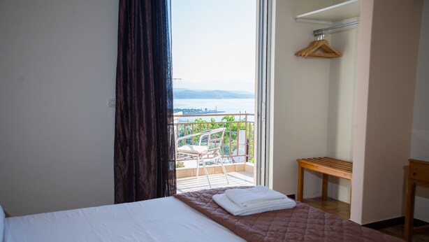 A hotel room with a bed, a wooden bench, a hanging coat rack, and a balcony with a view of the city and water. - Hotel Milton (East Mani)