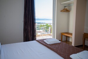 Habitación de hotel con cama, banco de madera, perchero y balcón con vistas a la ciudad y al agua.