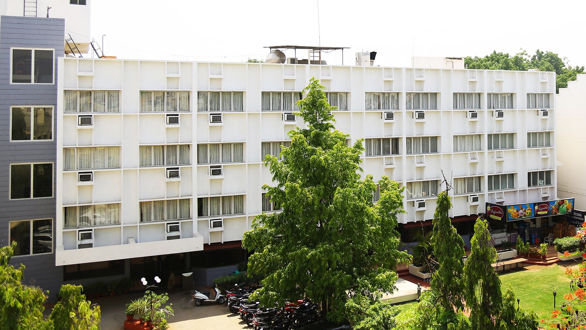 Et moderne hotel med flere etager, der har airconditionanlæg, en antenne på taget og en gårdhave med parkerede scootere og plantekasser.