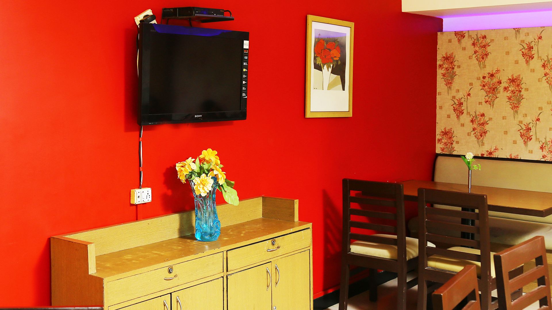 A room with a red wall, a wooden cabinet with a vase of flowers, a television mounted above it, and a framed picture on the wall.