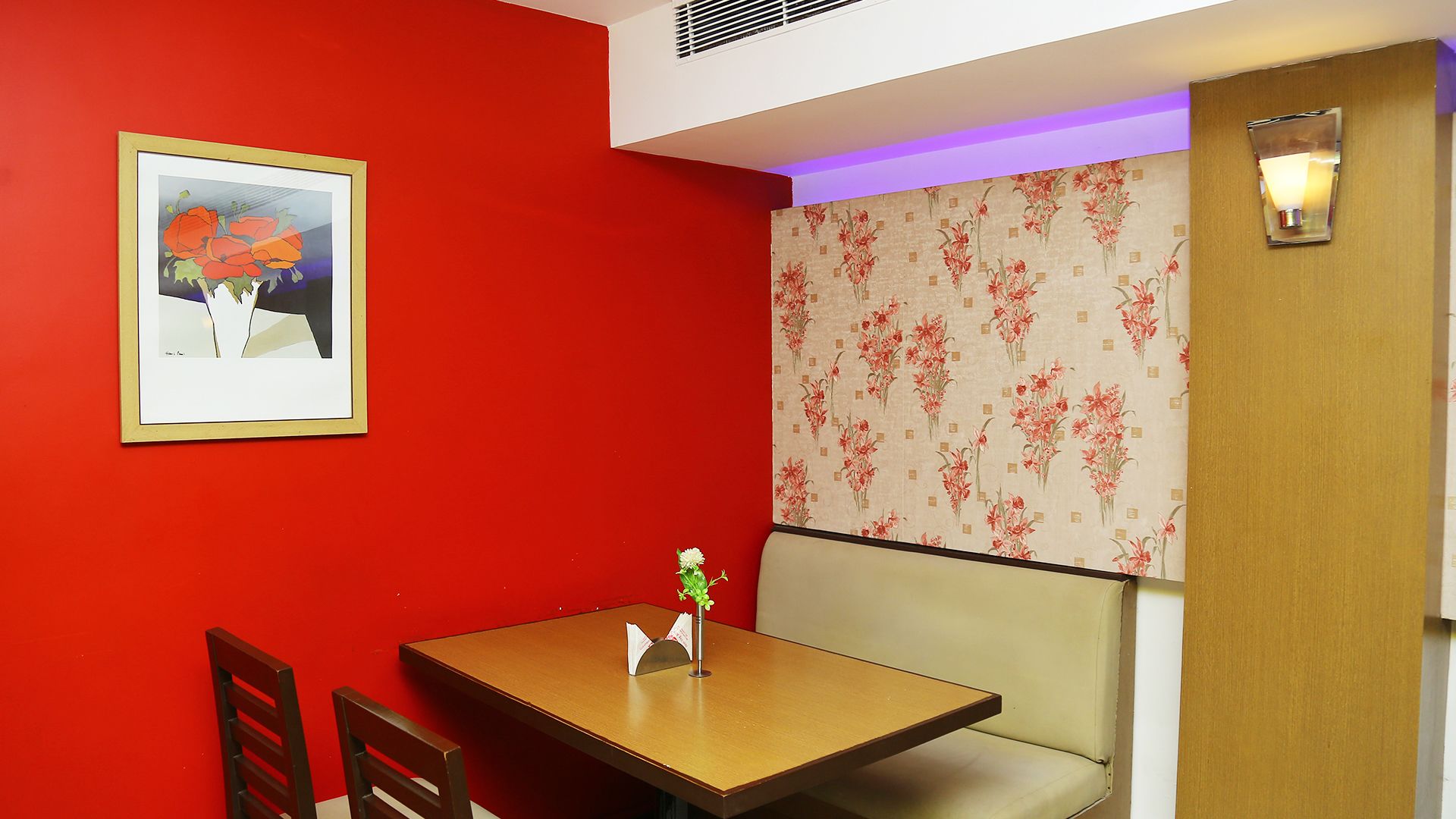 A dining area with a red wall, a framed picture, a floral wallpaper, a table, and a chair.
