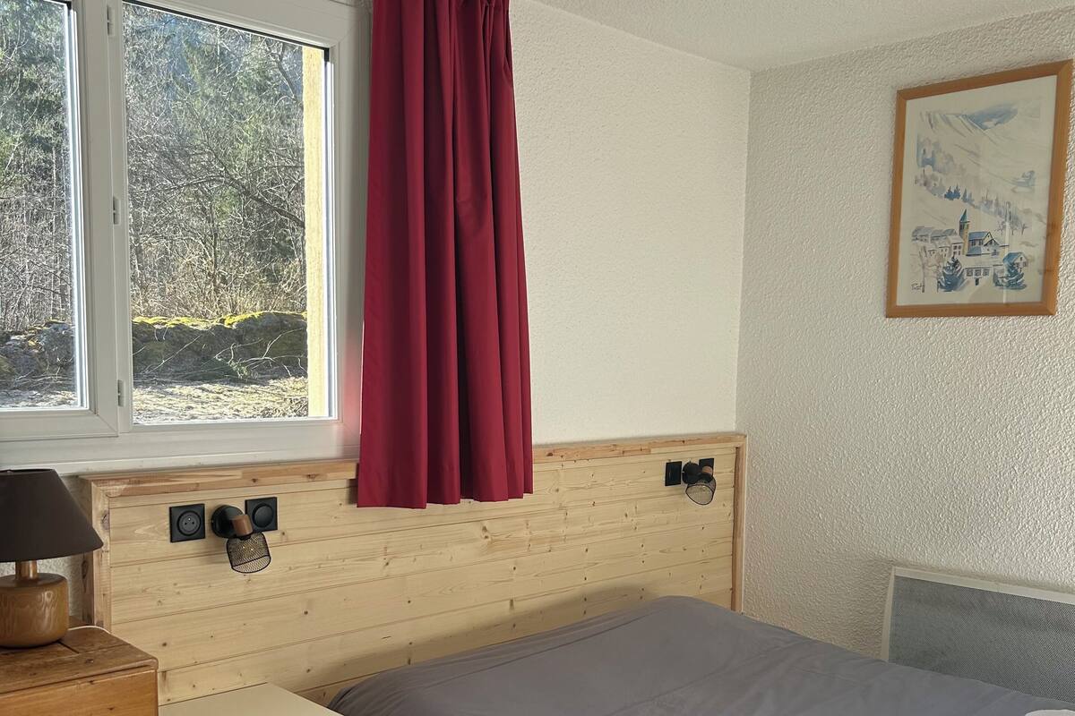A bedroom with a bed, a wooden dresser, a nightstand, a window with curtains, a radiator, and a wall-mounted picture.