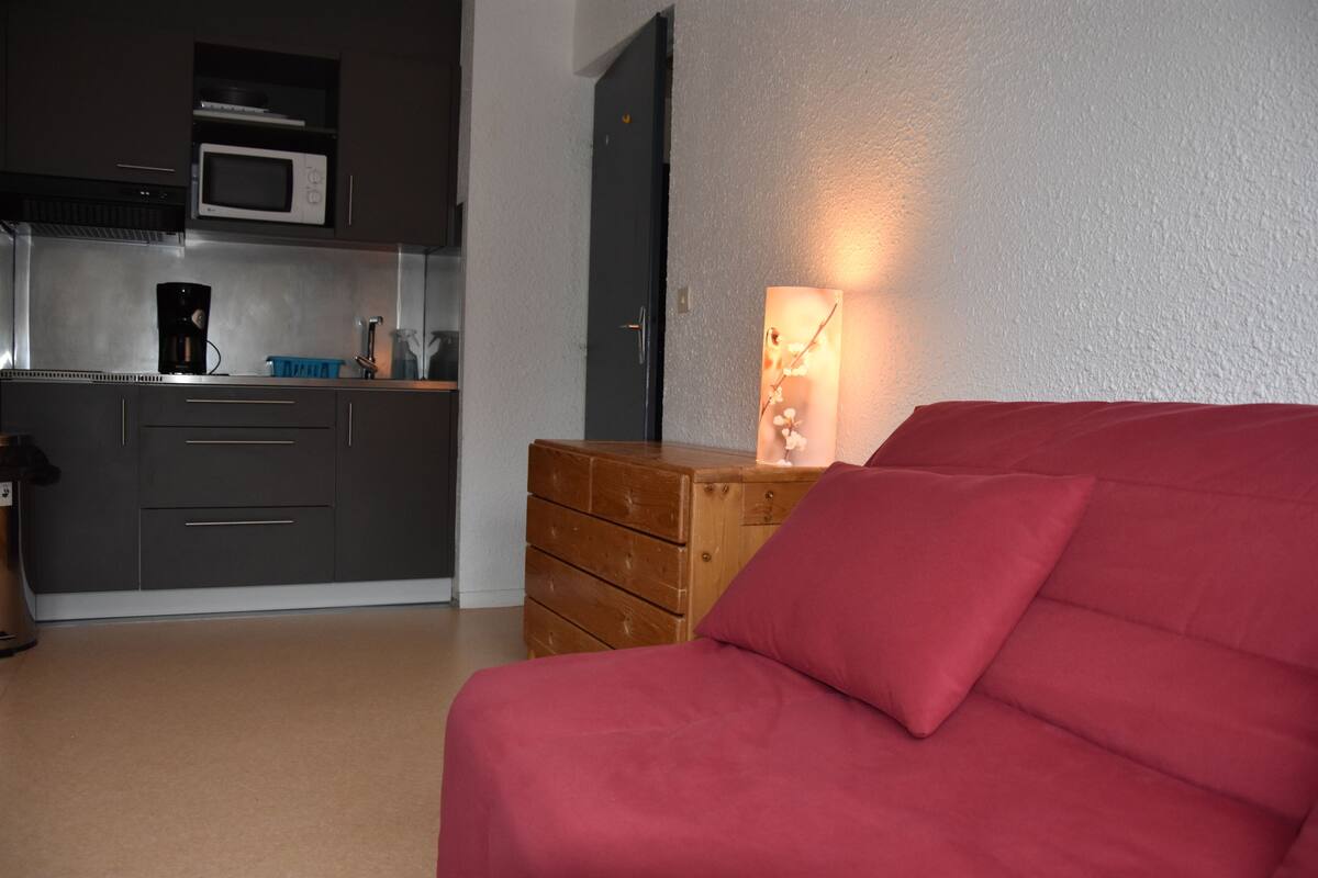 A bedroom with a red bed, a wooden dresser, a microwave in the kitchen area, and a wall-mounted light fixture.
