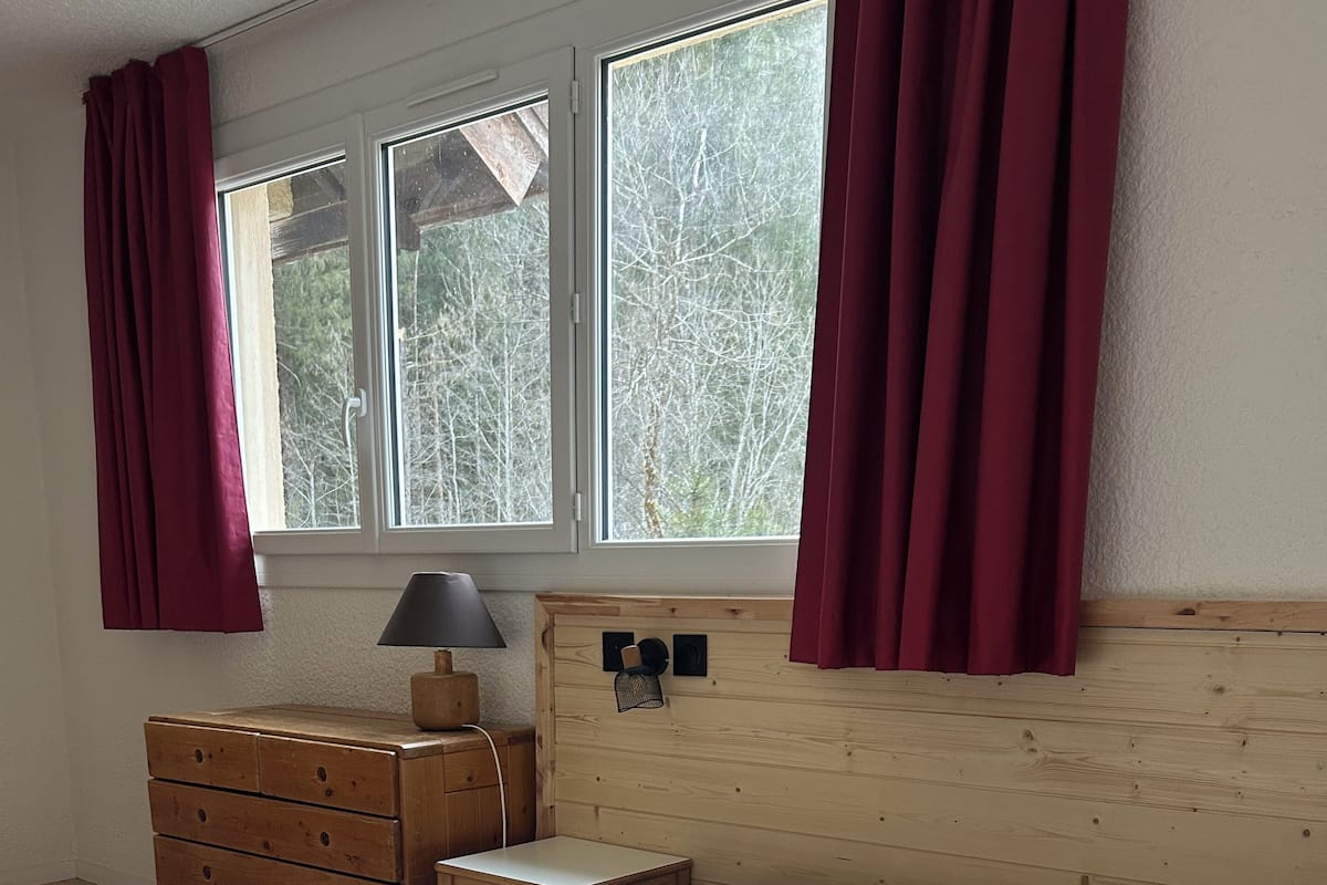 A room with a bed, wooden dresser, bedside table, lamp, and a window with curtains.