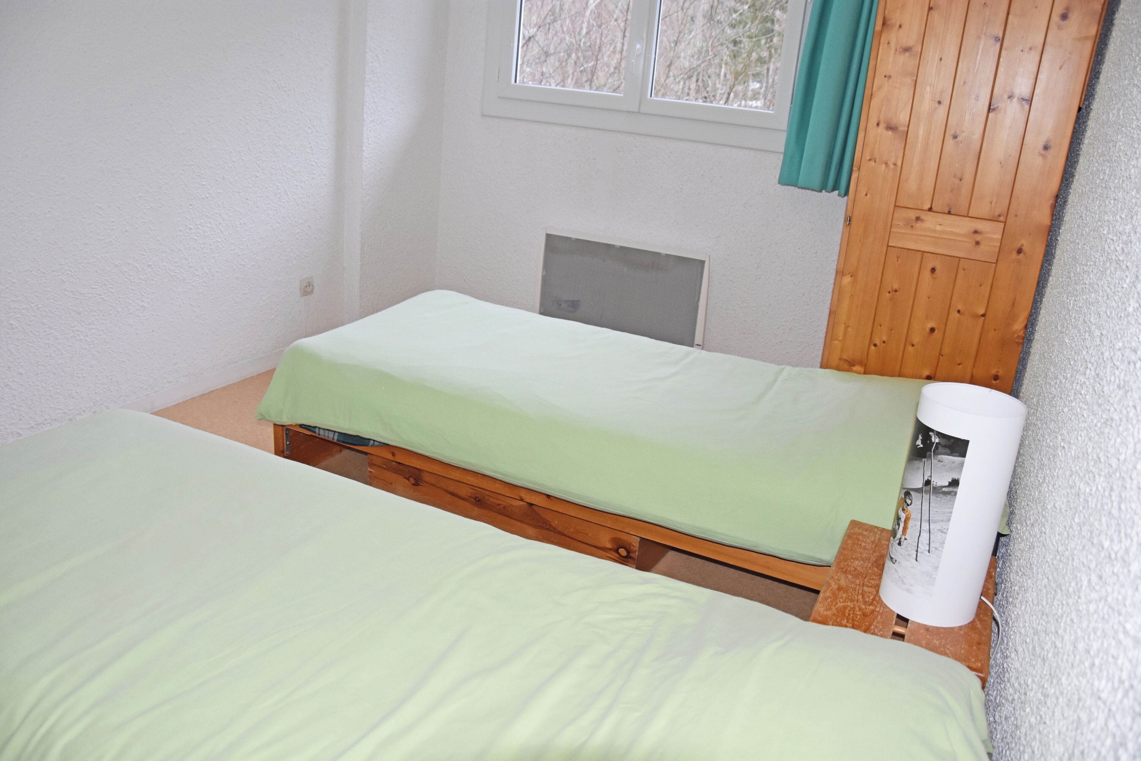 Una habitación con dos camas, una ventana con cortinas, un armario de madera y un pequeño calentador.