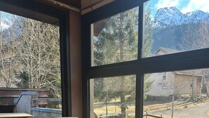 A room with a grey sofa, a round wooden table, and a large window with a view of a mountain and trees. - Village vacances Cévéo du Haut Breda (Le Haut-Bréda)