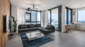 A modern living room with a sectional sofa, a coffee table, and a view of the city through large windows. - Front of Beach next to RoyalBeach Hotel (Tel Aviv)