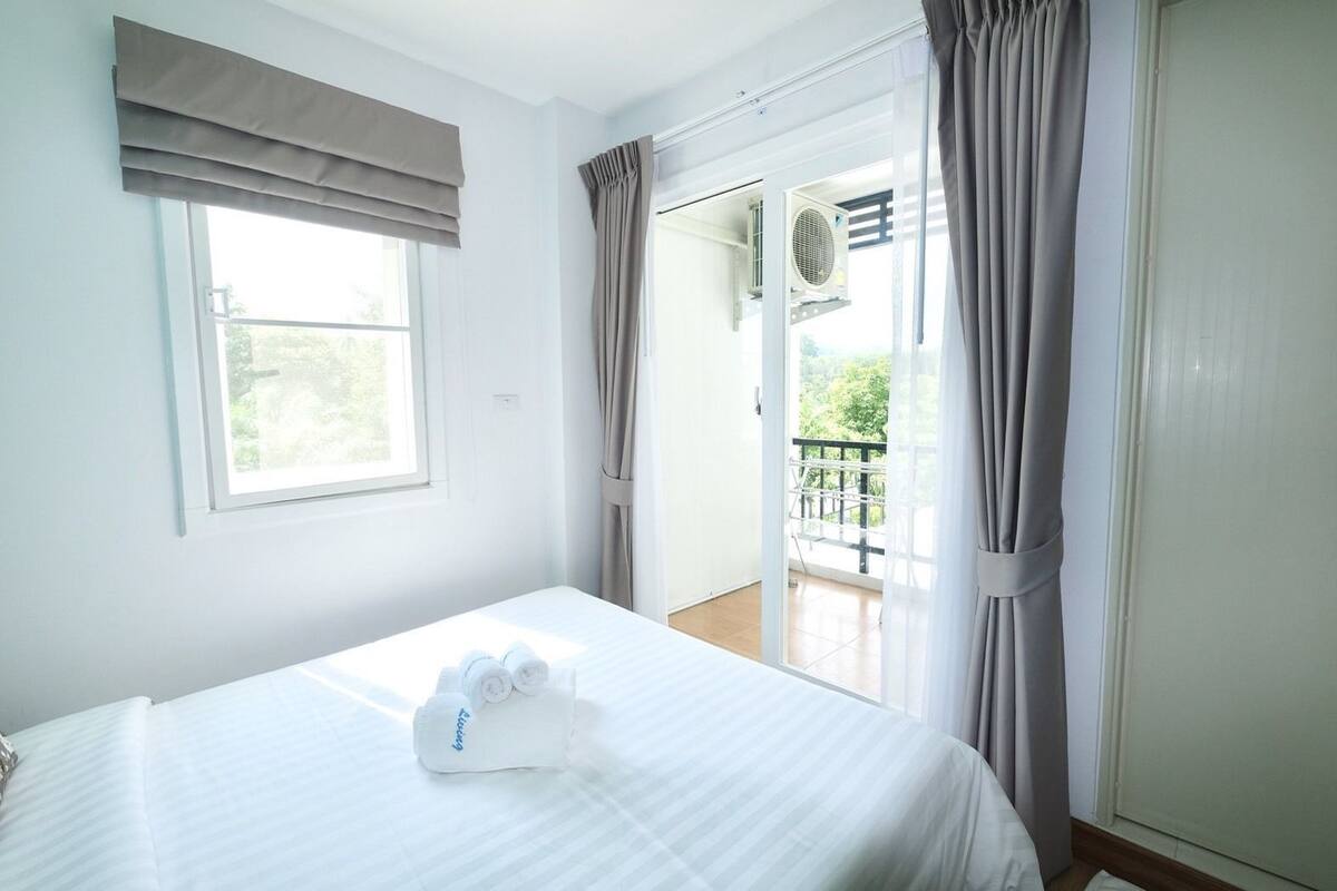 A hotel room with a bed, a window with curtains, a balcony with a view, and an air conditioning unit.