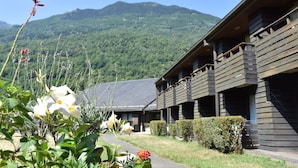 A wooden building with a mountain backdrop, a paved pathway, and a variety of plants in the foreground. - Village Cévéo de Luz-Saint-Sauveur (Luz-Saint-Sauveur)