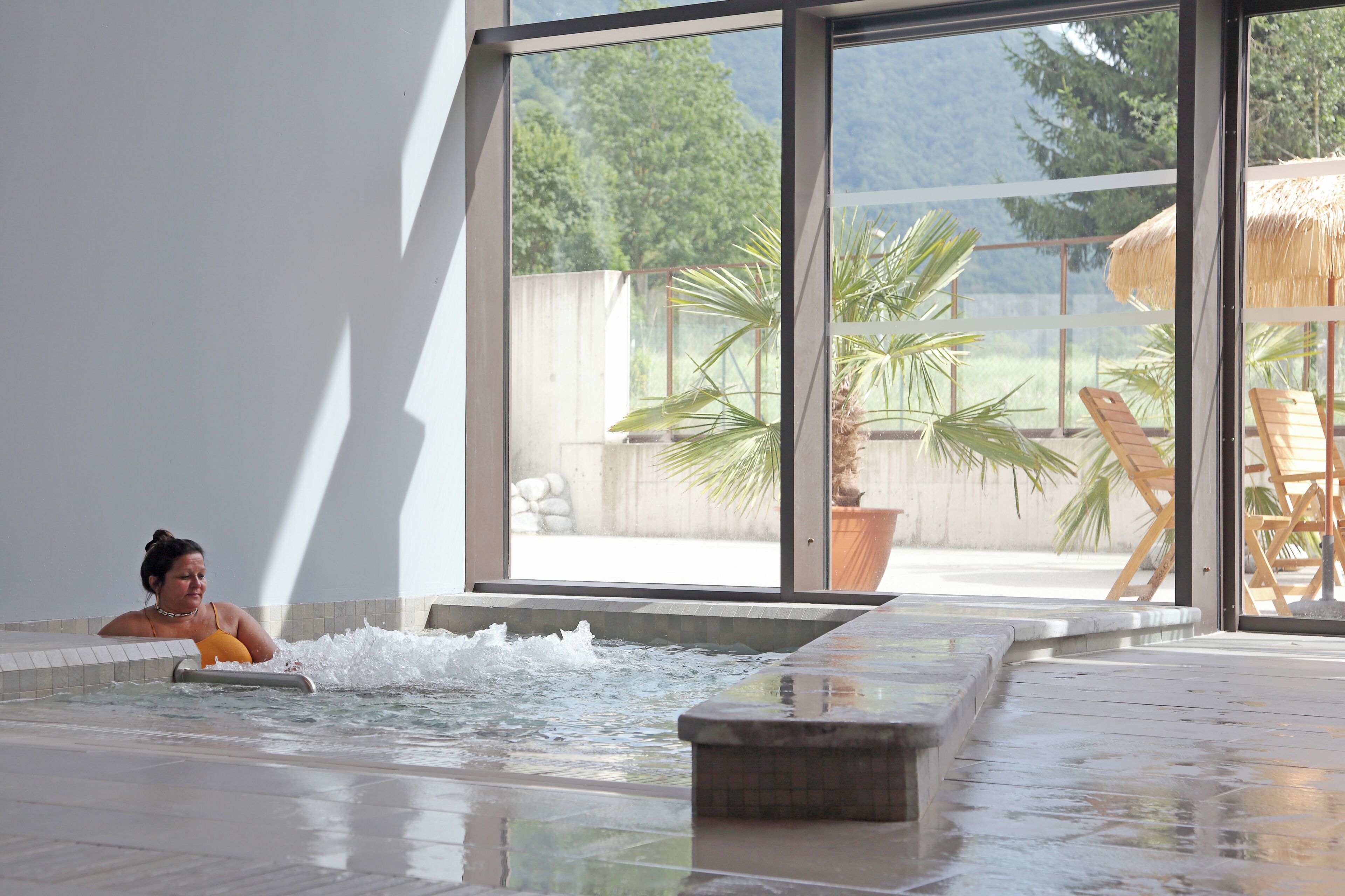 Une personne se trouve dans un bain à remous, avec vue sur un espace extérieur aménagé avec des chaises et un parasol en chaume.