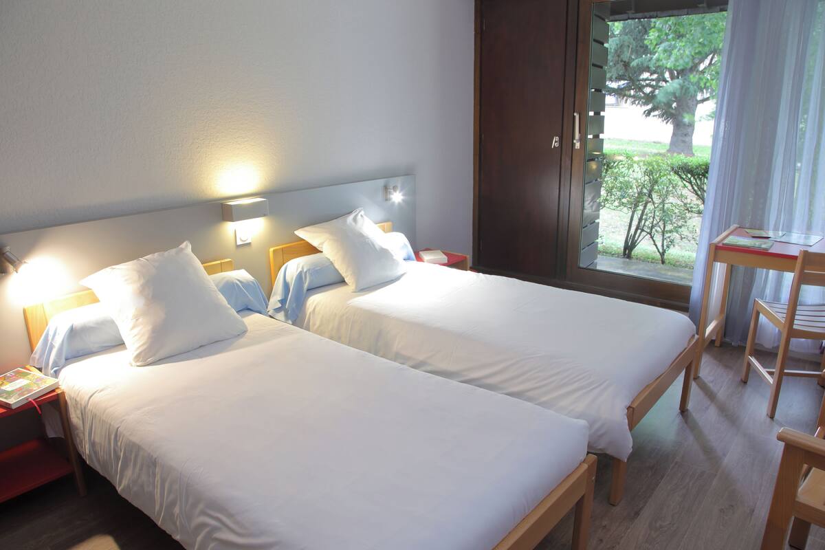 A hotel room with two beds, a desk, and a view of the outdoors.
