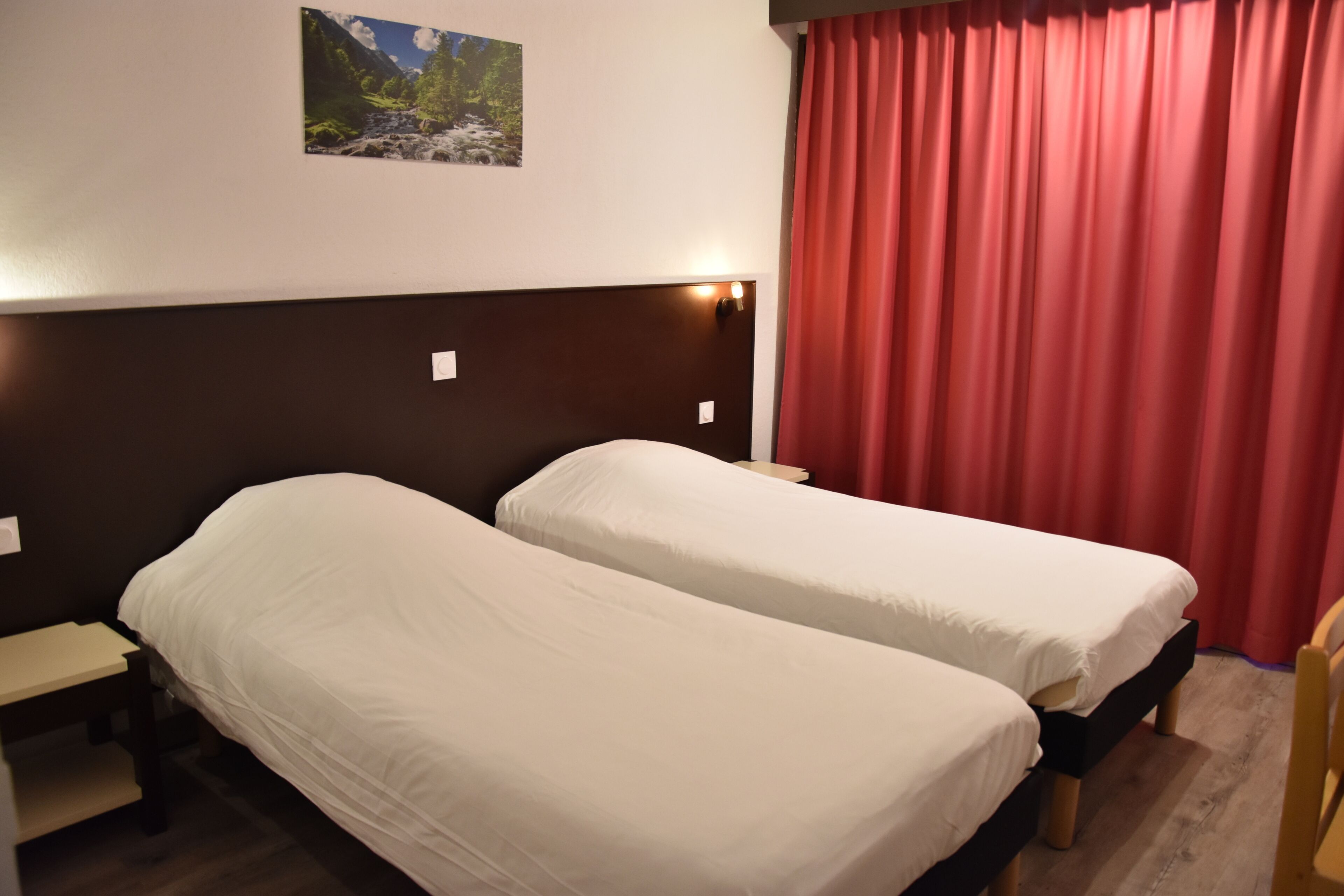 A room with two single beds, a nightstand, a red curtain, and a wall-mounted picture.