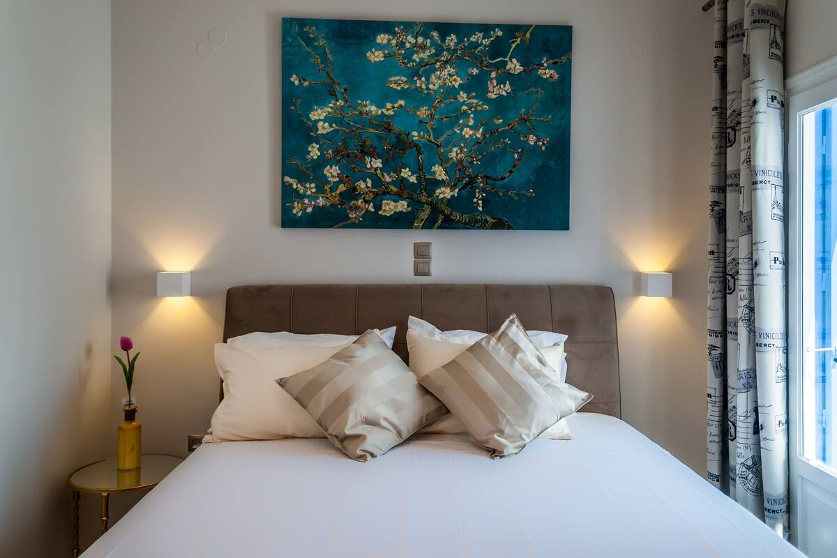 A neatly made bed with two pillows, a wall-mounted painting of a tree, and a window with curtains.