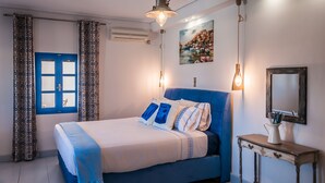 Un dormitori amb un llit blau, cortines estampades, un quadre, una taula de nit i un mirall.