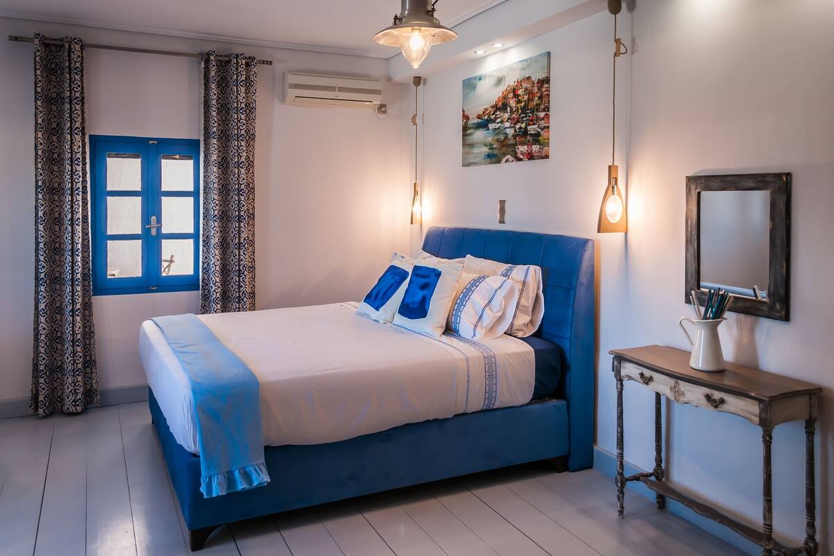 A bedroom with a blue bed, patterned curtains, a painting, a bedside table, and a mirror.