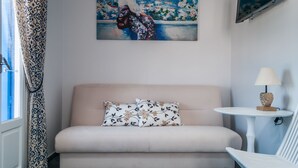 Un sofà beix amb coixins florals, una taula petita amb una làmpada, i un quadre a la paret.
