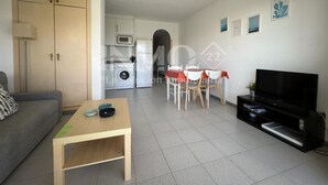 Una sala de estar con un sofá, una mesa de centro, un televisor y una zona de comedor con sillas y una mesa. Se observa una cocina al fondo con una lavadora y un microondas.