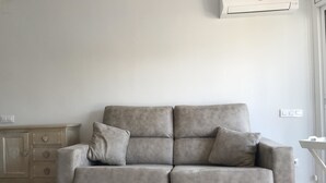 A grey sofa with two cushions in a room with a wooden cabinet and a white air conditioning unit on the wall. - Estudio Reformado Regueral - 123A (Cambrils)