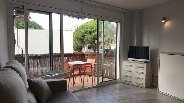A modern living room with a grey sofa, a TV on a cabinet, and a sliding glass door leading to an outdoor patio with a table and chairs. - Estudio Reformado Regueral - 123A (Cambrils)