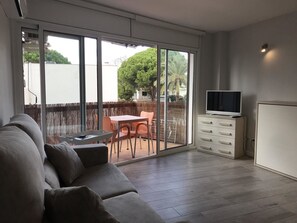 A modern living room with a grey sofa, a TV on a cabinet, and a sliding glass door leading to an outdoor patio with a table and chairs. - Estudio Reformado Regueral - 123A (Cambrils)