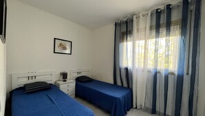 A bedroom with two single beds, a bedside table with a lamp, a framed picture on the wall, and a window with curtains. - Apartamento Cambrils Park - 104B (Cambrils)