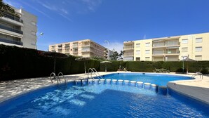 A swimming pool with a tiled edge, surrounded by a hedge and a building in the background. - Apartamento Cambrils Park - 104B (Cambrils)