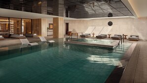 A modern indoor swimming pool with lounge areas, a ceiling with a geometric design, and a clock on the wall.