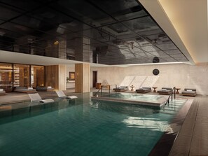 A modern indoor swimming pool with lounge areas, a ceiling with a geometric design, and a clock on the wall.