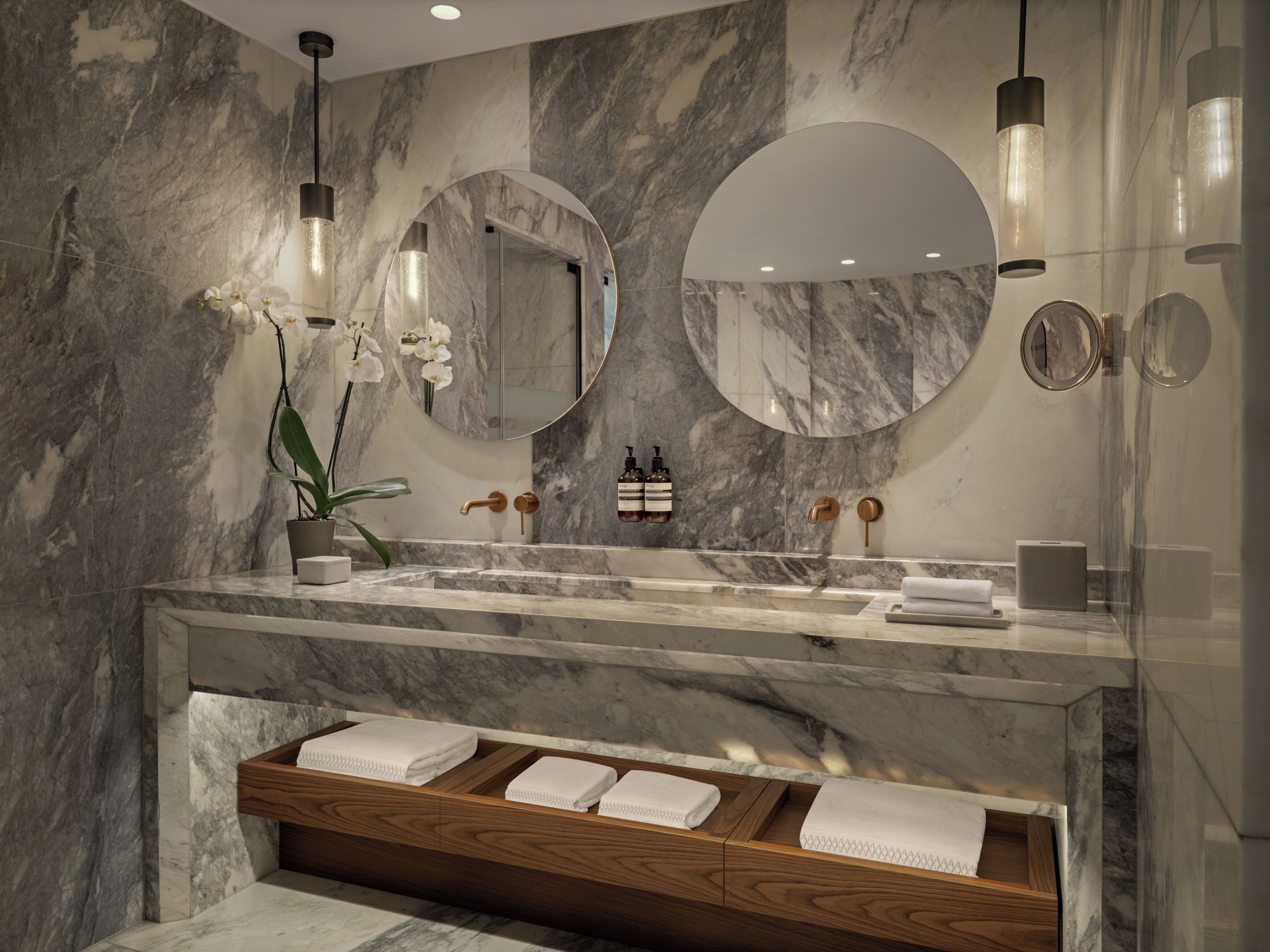 A modern bathroom with marble walls, a large sink, two mirrors, and a wooden vanity with towels.