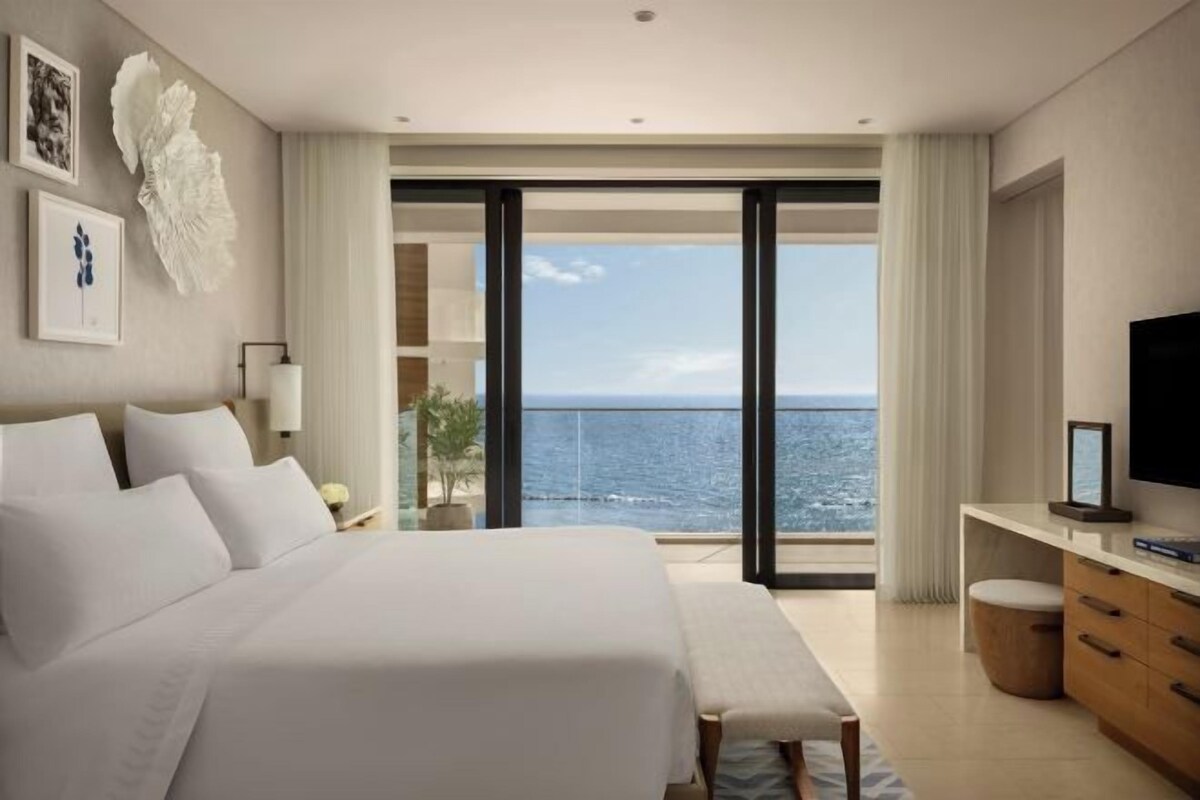 A bedroom with a large bed, a desk, and a television. There is a balcony with a view of the ocean.