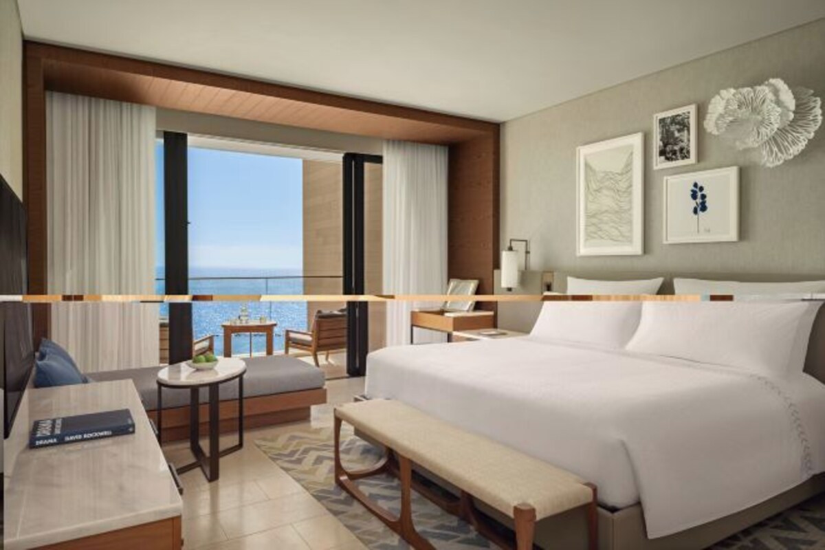A hotel room with a large bed, a desk, a chair, a small table, and a bench. There is a view of the ocean through large windows.