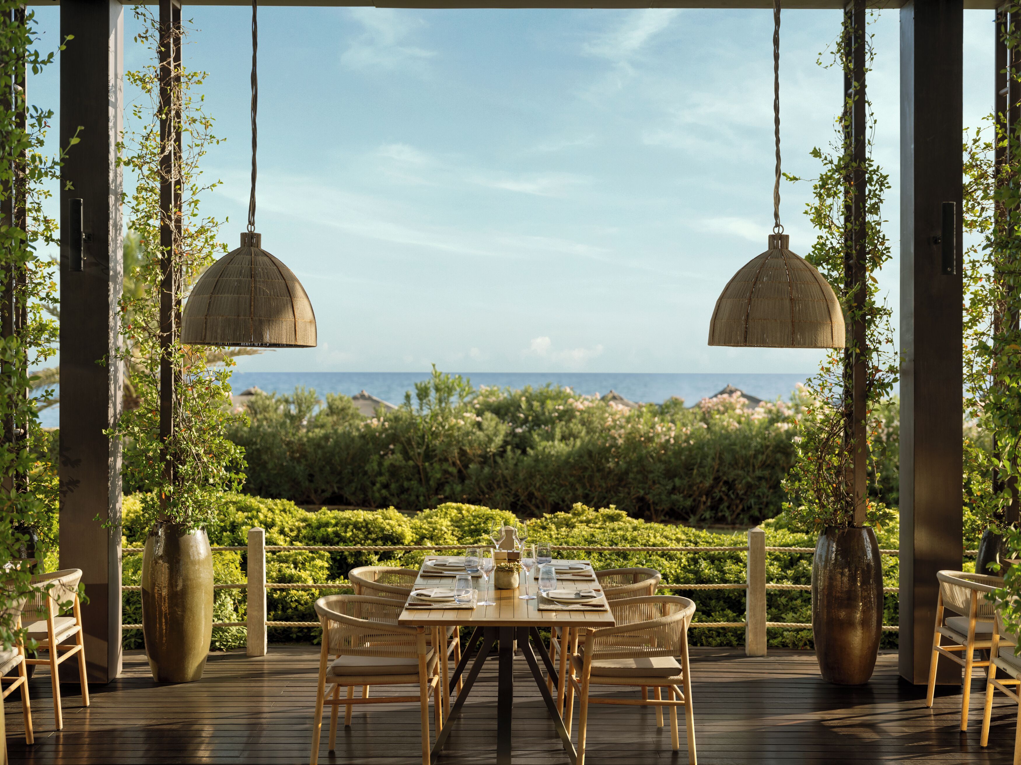 A wooden deck with a table set for dining, surrounded by chairs, and overlooking a lush garden and the sea.