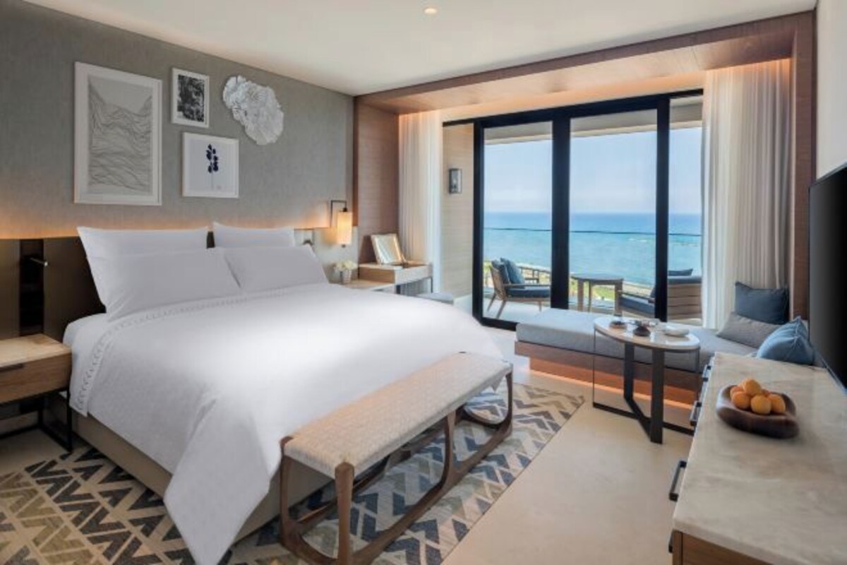 A modern hotel room with a large bed, a small table, a bench, and a view of the sea.