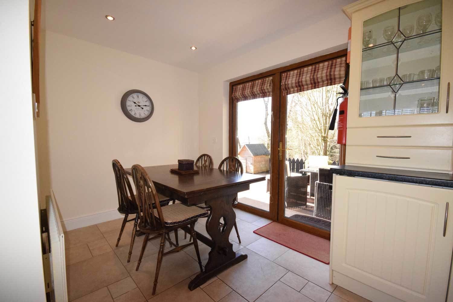 A dining area with a wooden table and chairs, a clock on the wall, and a kitchen with cabinets and a glass display case.