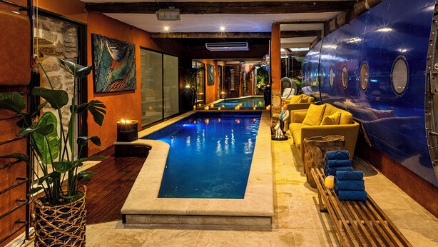 Un área de piscina con borde de piedra, pared azul con ventanas circulares y un sofá amarillo con cojines.