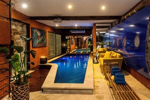 Un área de piscina con borde de piedra, pared azul con ventanas circulares y un sofá amarillo con cojines.